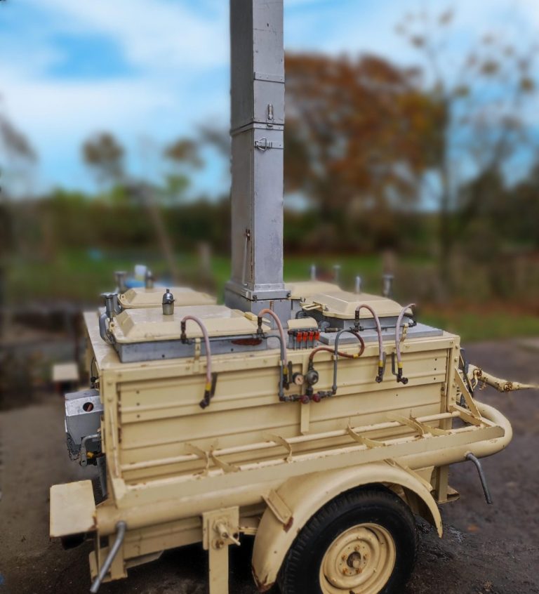 Gelbes Fahrzeug mit hohem Auspuffrohr, auf einem Schotterboden, umgeben von Natur.